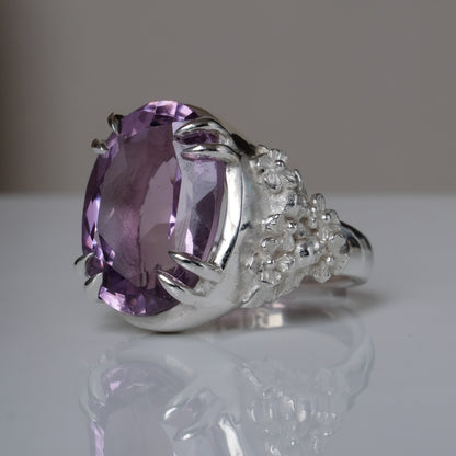 Ring claw set with large amethyst gemstone with skull and floral detail on side.  made in sterling silver on reflective surface