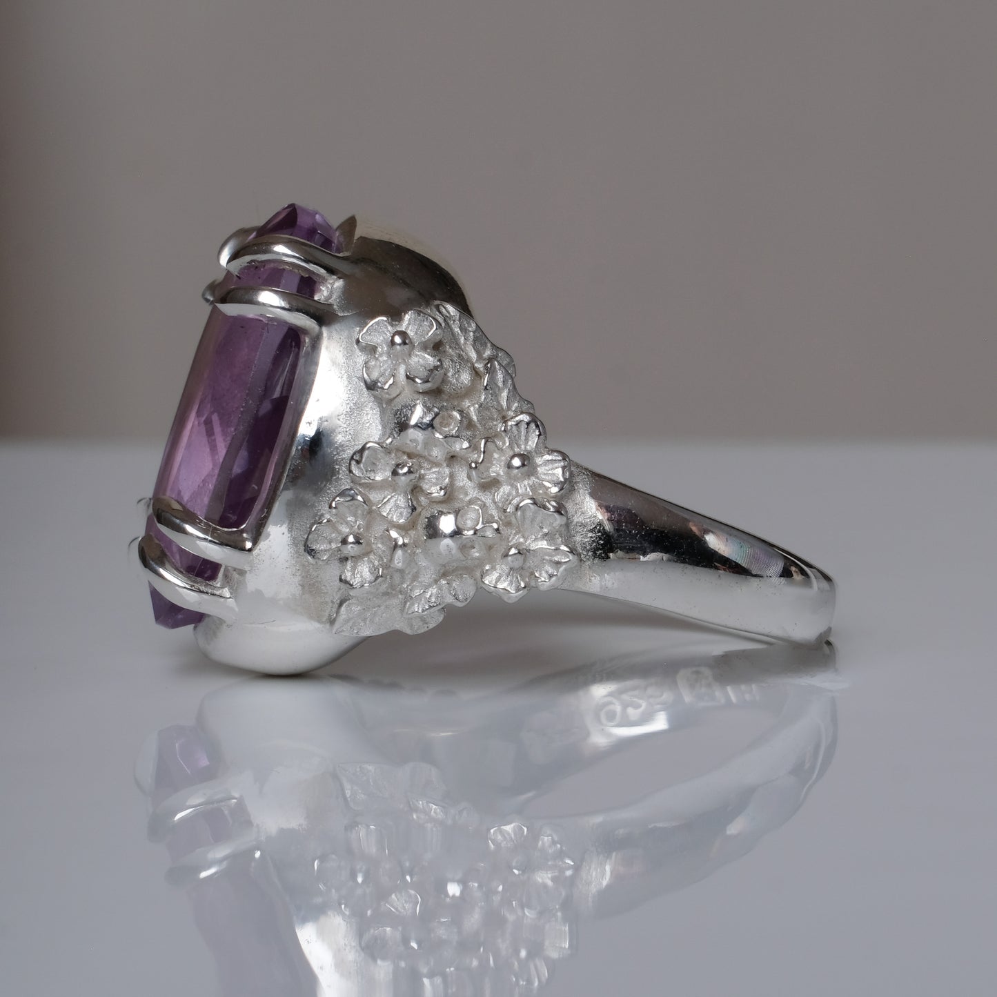 Ring claw set with large amethyst gemstone with skull and floral detail on side.  made in sterling silver on reflective surface