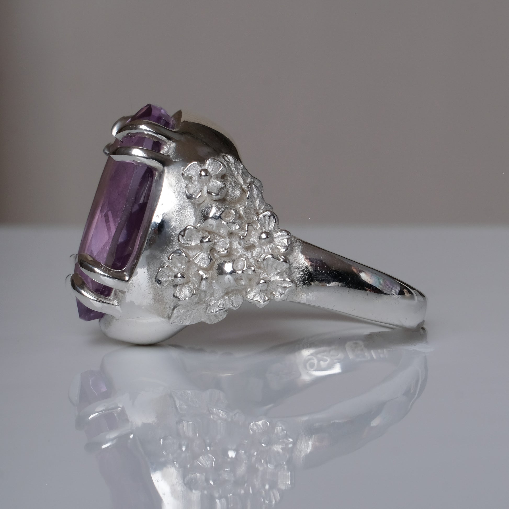 Ring claw set with large amethyst gemstone with skull and floral detail on side.  made in sterling silver on reflective surface