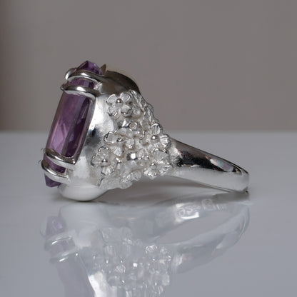Ring claw set with large amethyst gemstone with skull and floral detail on side.  made in sterling silver on reflective surface