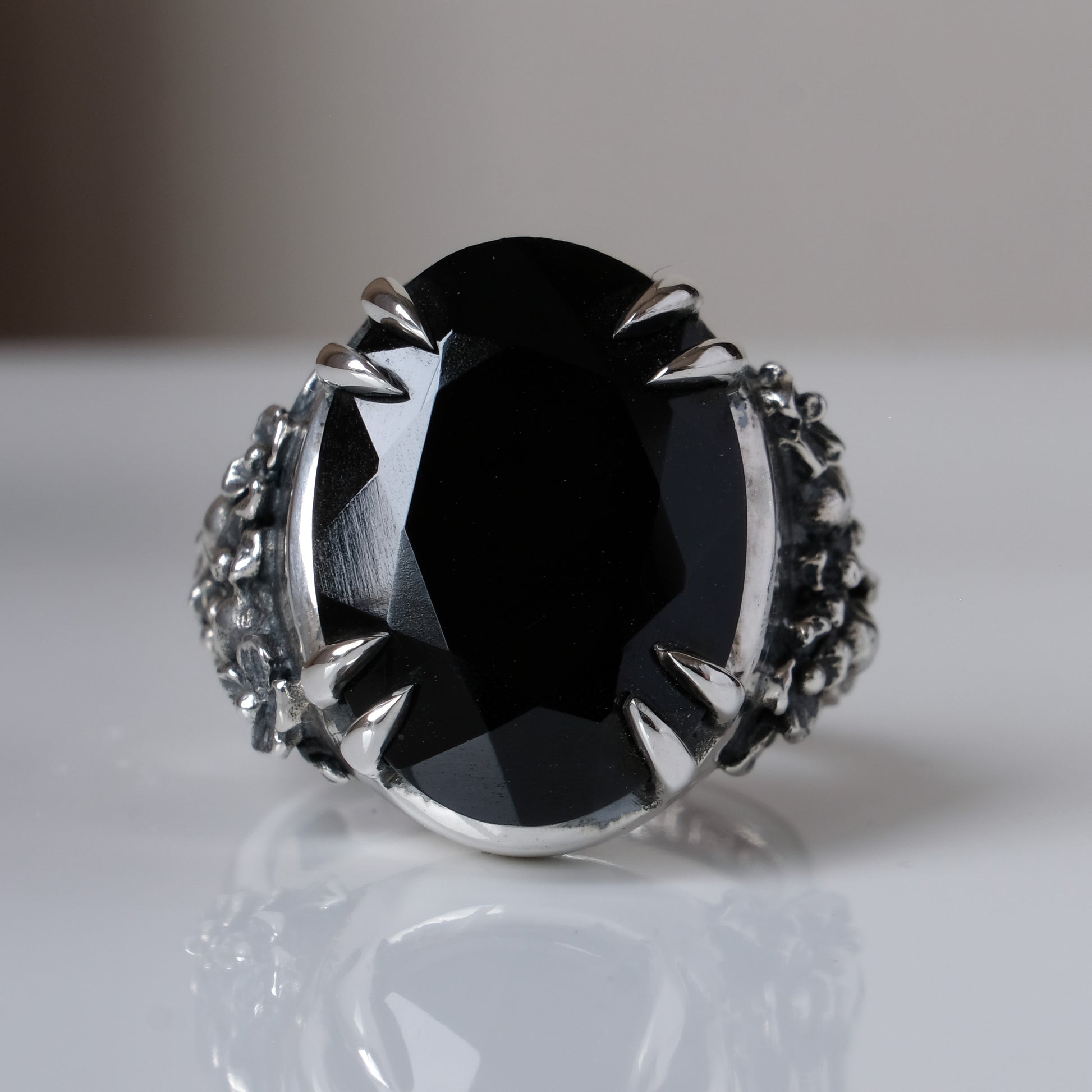 Ring claw set with large black spinel gemstone with skull and floral detail on side.  made in sterling silver on reflective surface