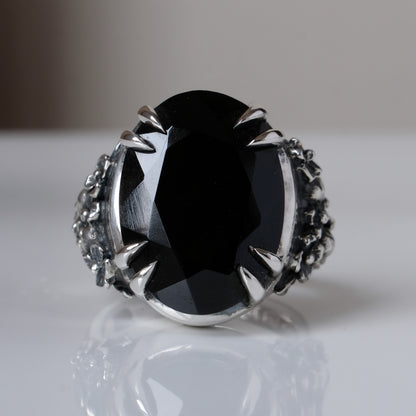 Ring claw set with large black spinel gemstone with skull and floral detail on side.  made in sterling silver on reflective surface