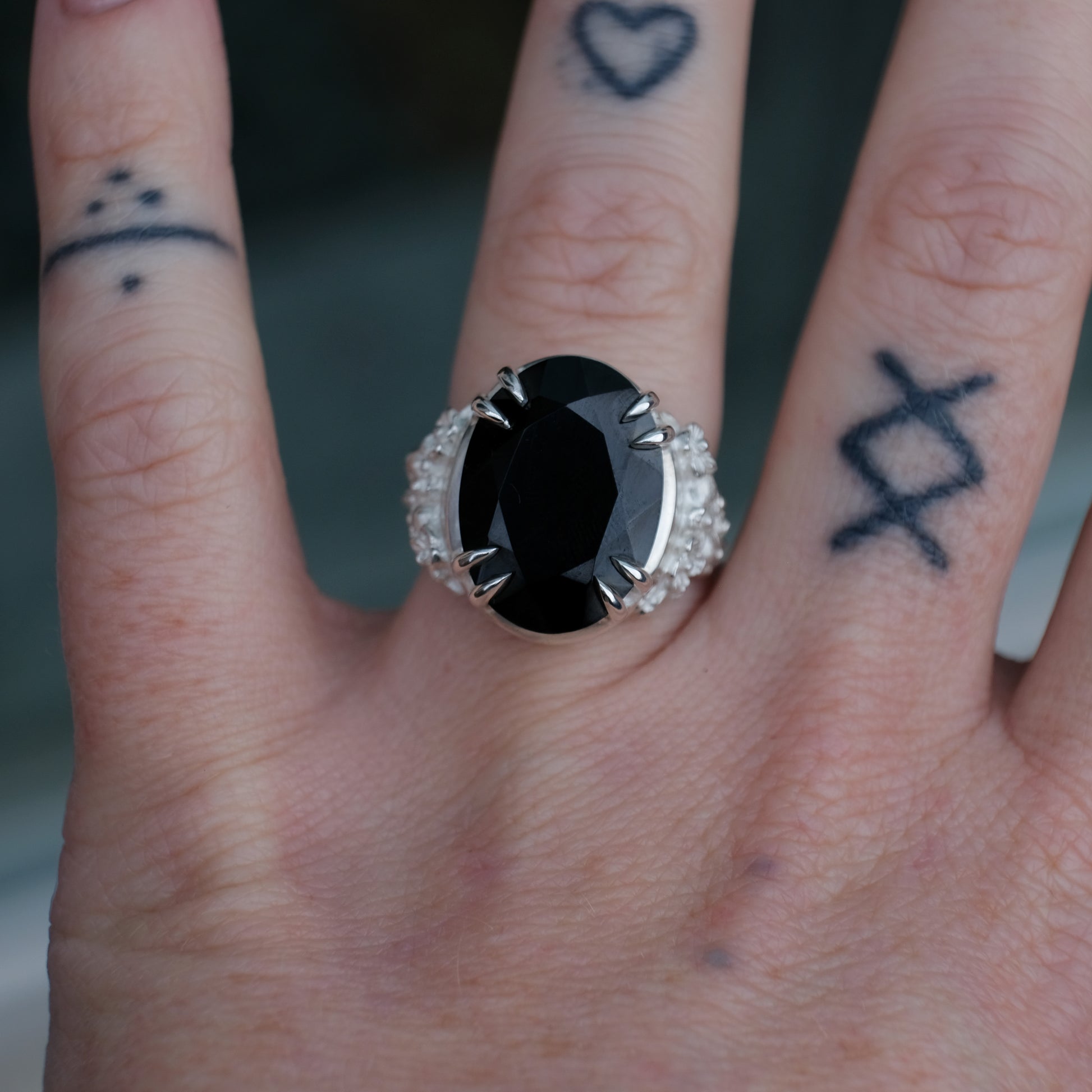 Ring claw set with large black spinel gemstone with skull and floral detail on side.  made in sterling silver worn on hand with tattoos