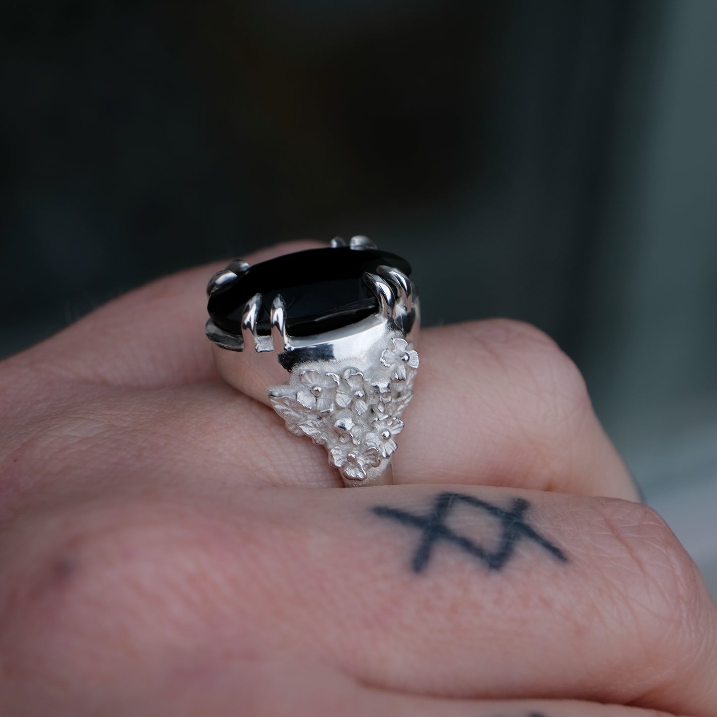 Ring claw set with large black spinel gemstone with skull and floral detail on side.  made in sterling silver worn on hand with tattoos
