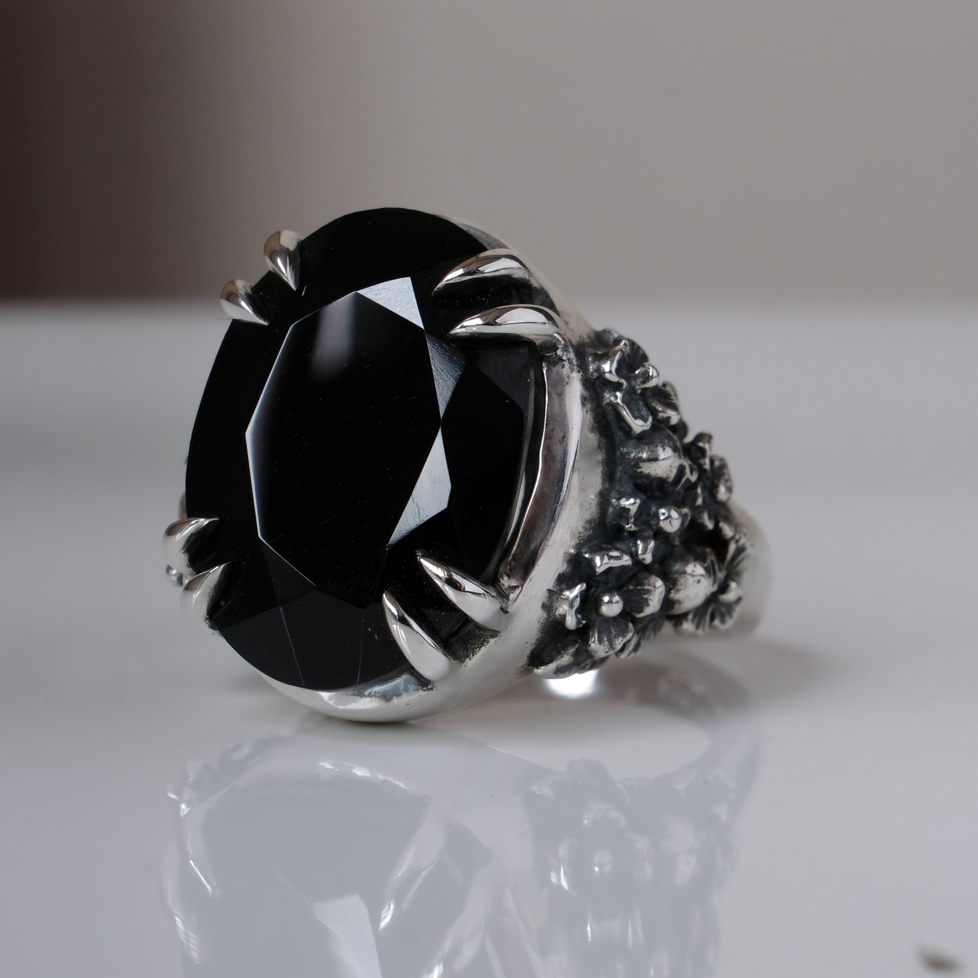Ring claw set with large black spinel gemstone with skull and floral detail on side.  made in sterling silver on reflective surface