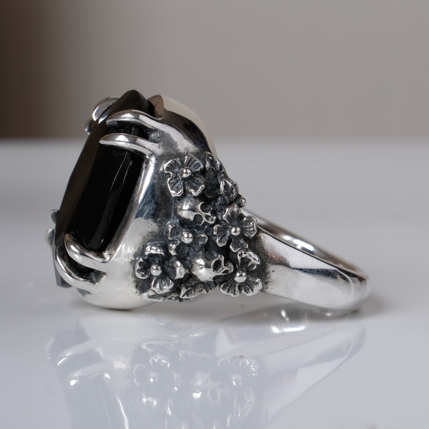 Ring claw set with large black spinel gemstone with skull and floral detail on side.  made in sterling silver on reflective surface