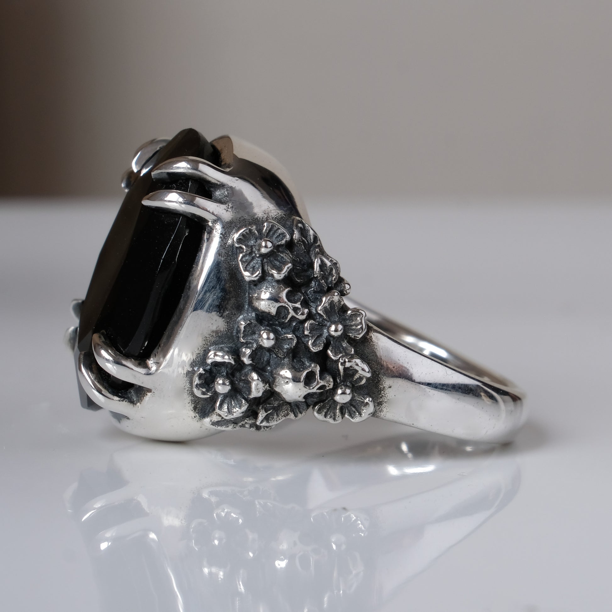 Ring claw set with large black spinel gemstone with skull and floral detail on side.  made in sterling silver on reflective surface