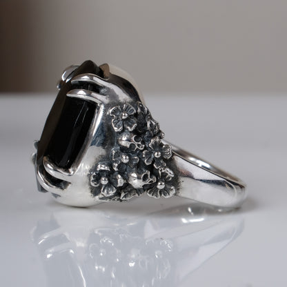 Ring claw set with large black spinel gemstone with skull and floral detail on side.  made in sterling silver on reflective surface
