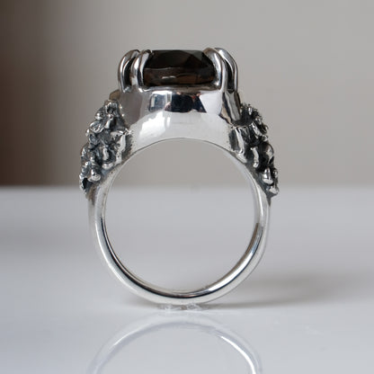 Ring claw set with large black spinel gemstone with skull and floral detail on side.  made in sterling silver on reflective surface