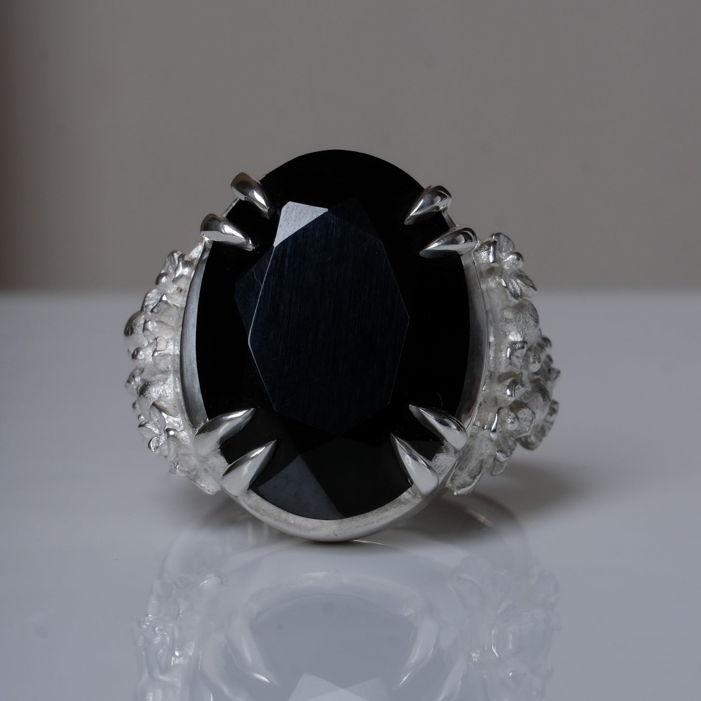 Ring claw set with large black spinel gemstone with skull and floral detail on side.  made in sterling silver on reflective surface