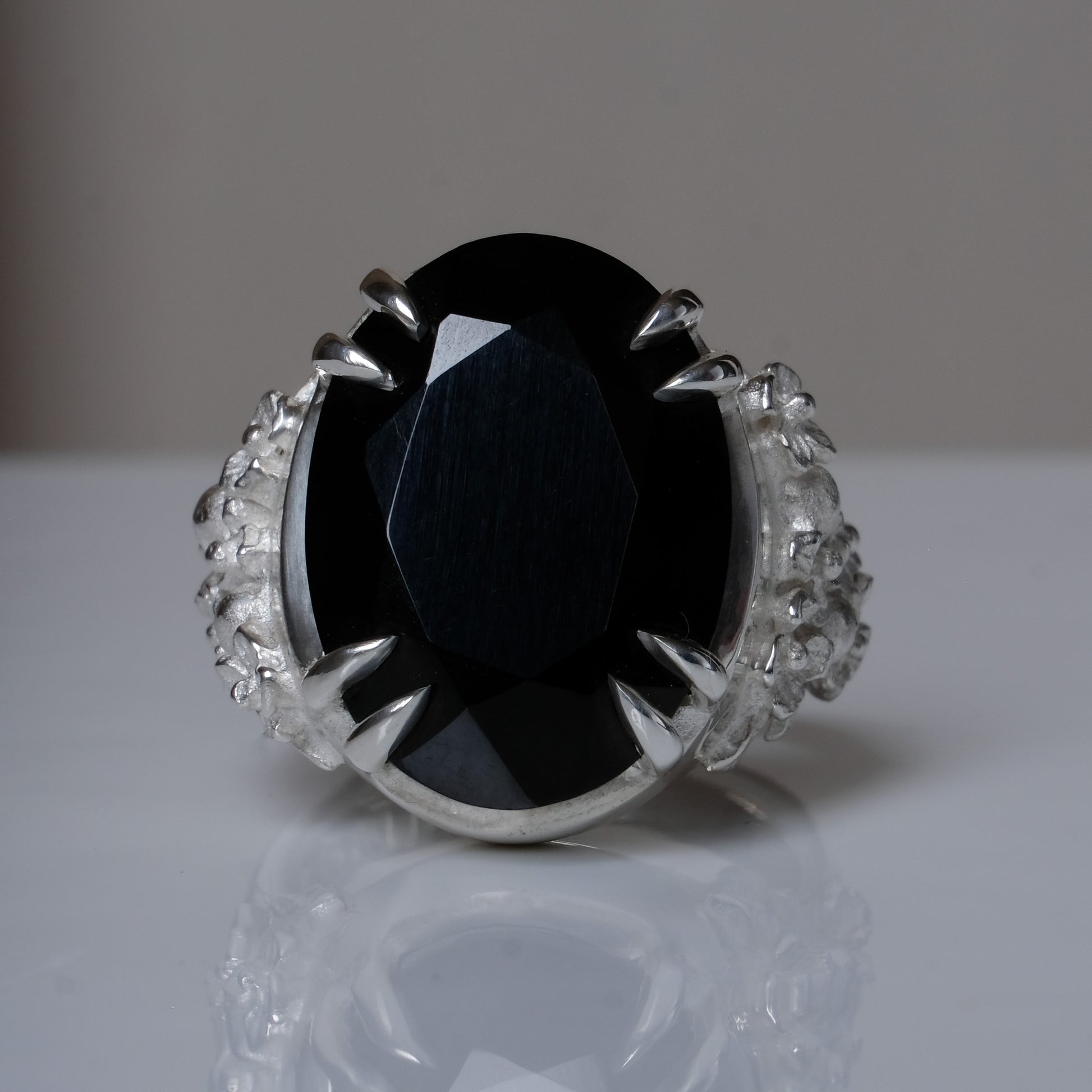 Ring claw set with large black spinel gemstone with skull and floral detail on side.  made in sterling silver on reflective surface