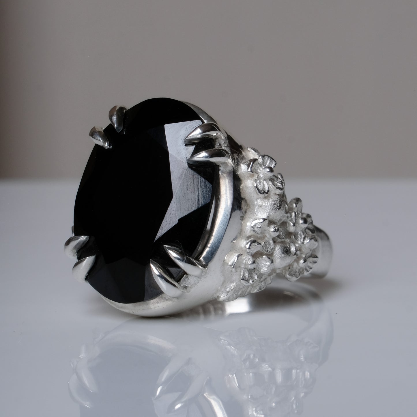 Ring claw set with large black spinel gemstone with skull and floral detail on side.  made in sterling silver on reflective surface