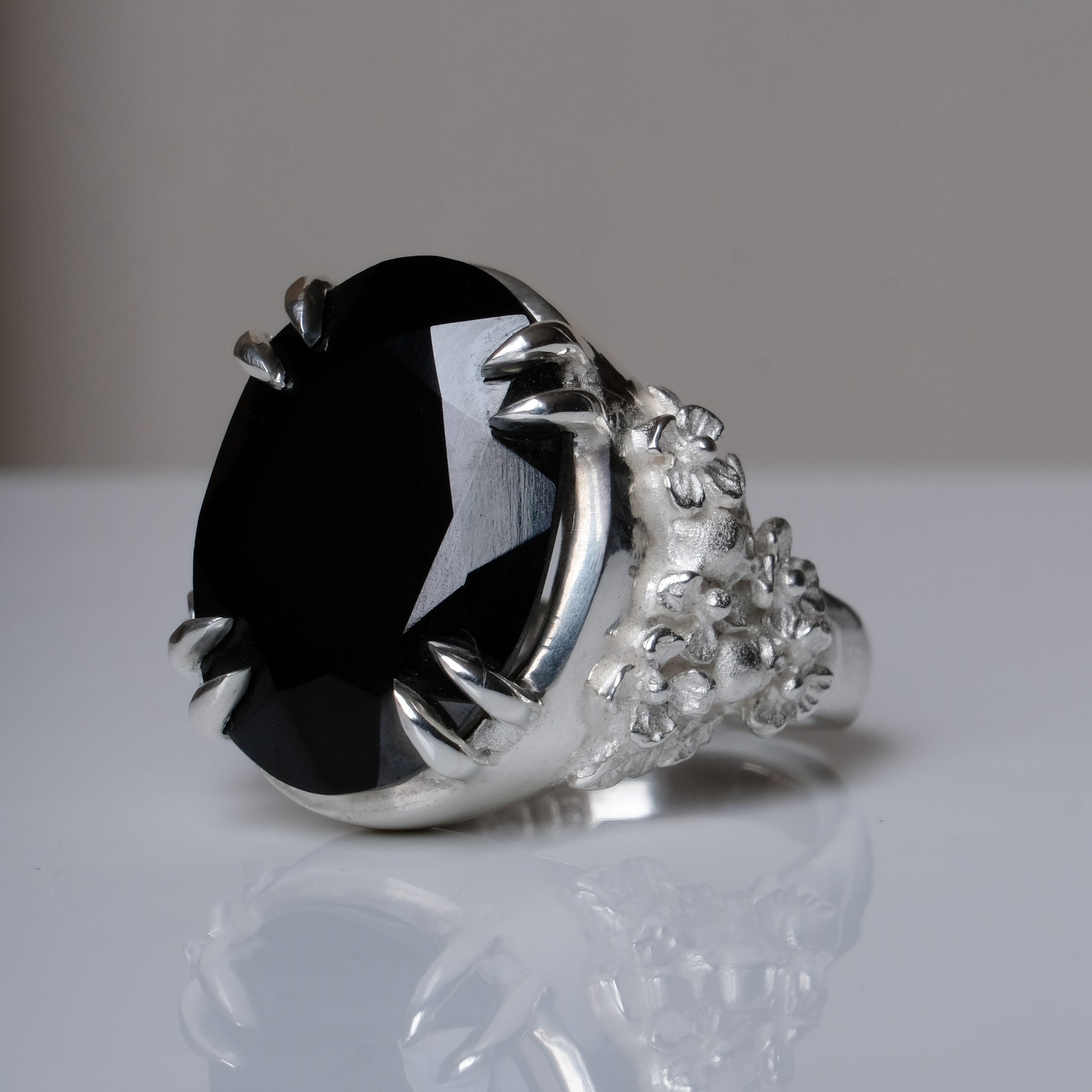 Ring claw set with large black spinel gemstone with skull and floral detail on side.  made in sterling silver on reflective surface
