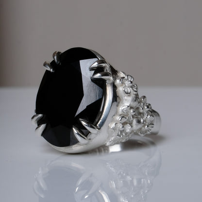 Ring claw set with large black spinel gemstone with skull and floral detail on side.  made in sterling silver on reflective surface