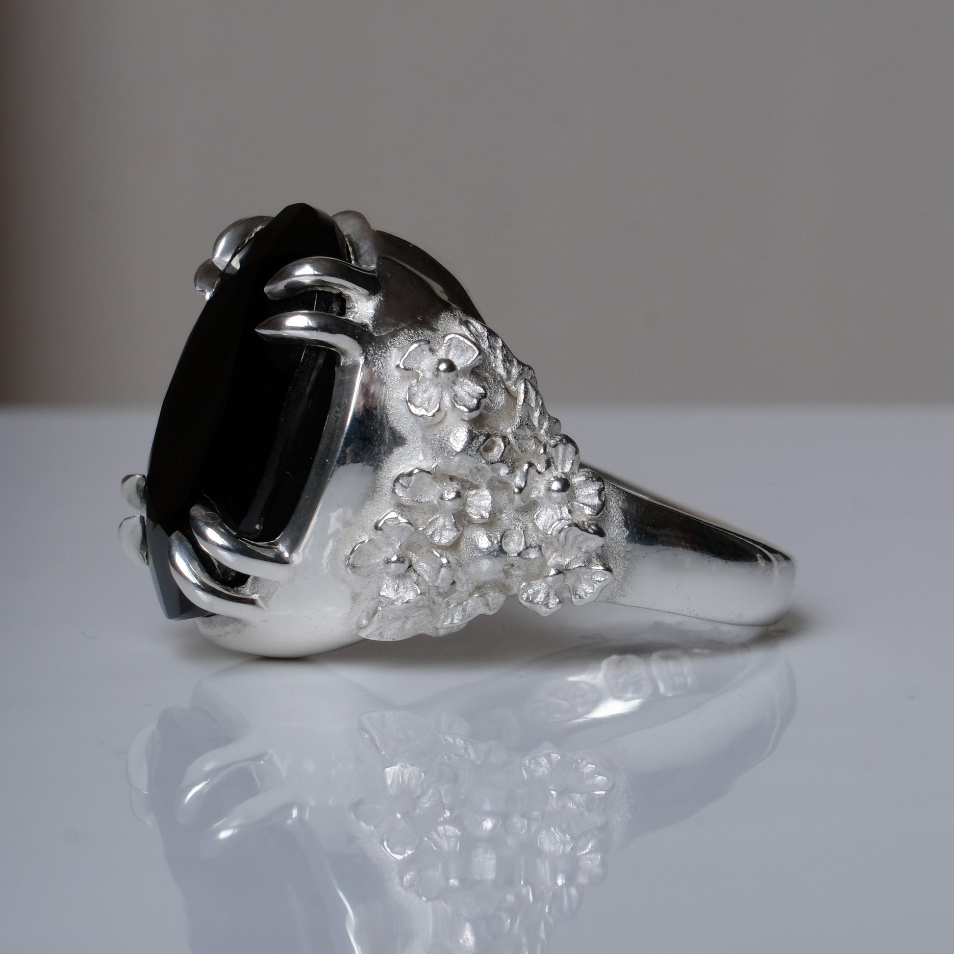 Ring claw set with large black spinel gemstone with skull and floral detail on side.  made in sterling silver on reflective surface