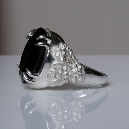 Ring claw set with large black spinel gemstone with skull and floral detail on side.  made in sterling silver on reflective surface