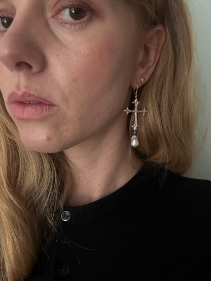 A close-up of a woman's ear wearing a silver-toned cross-shaped earring with a dangling pearl bead, against her blonde hair and black top by Charlie Shires