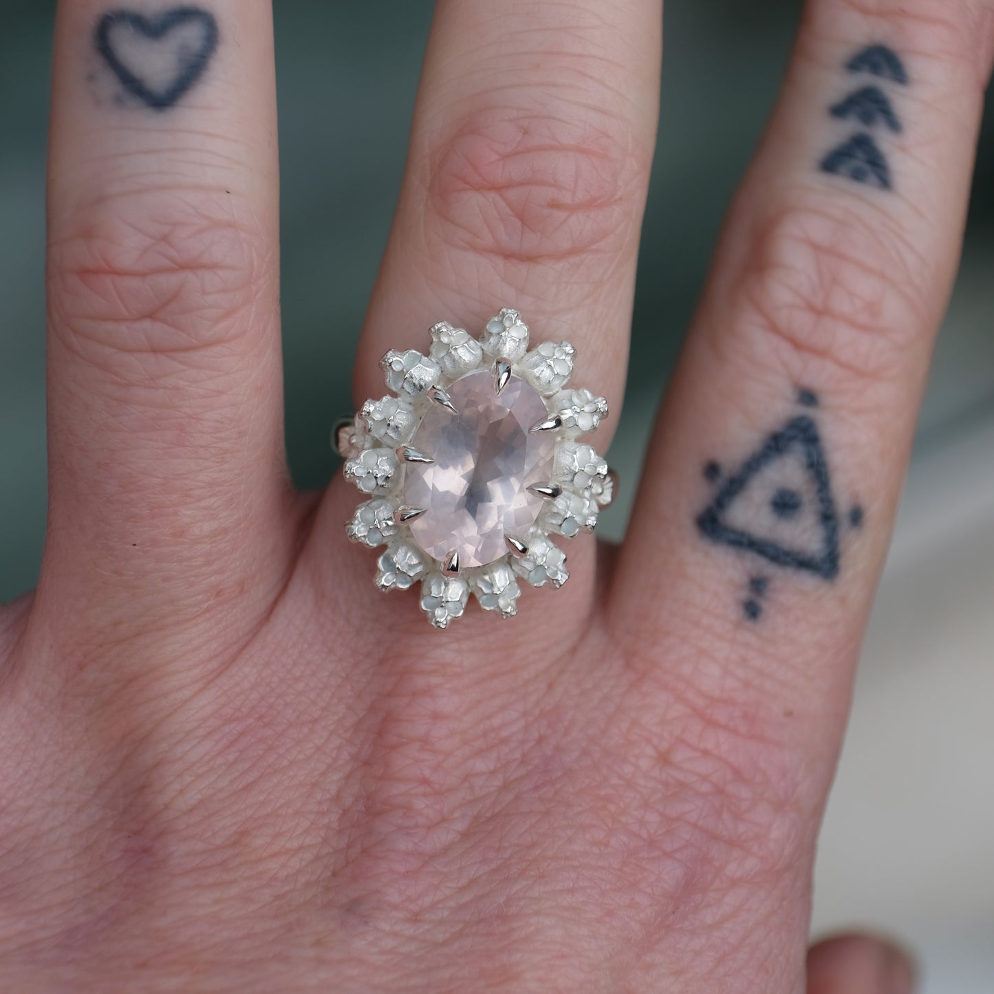 Ring claw set with rose quartz gemstone and silver skull halo design worn on hand with tattoos