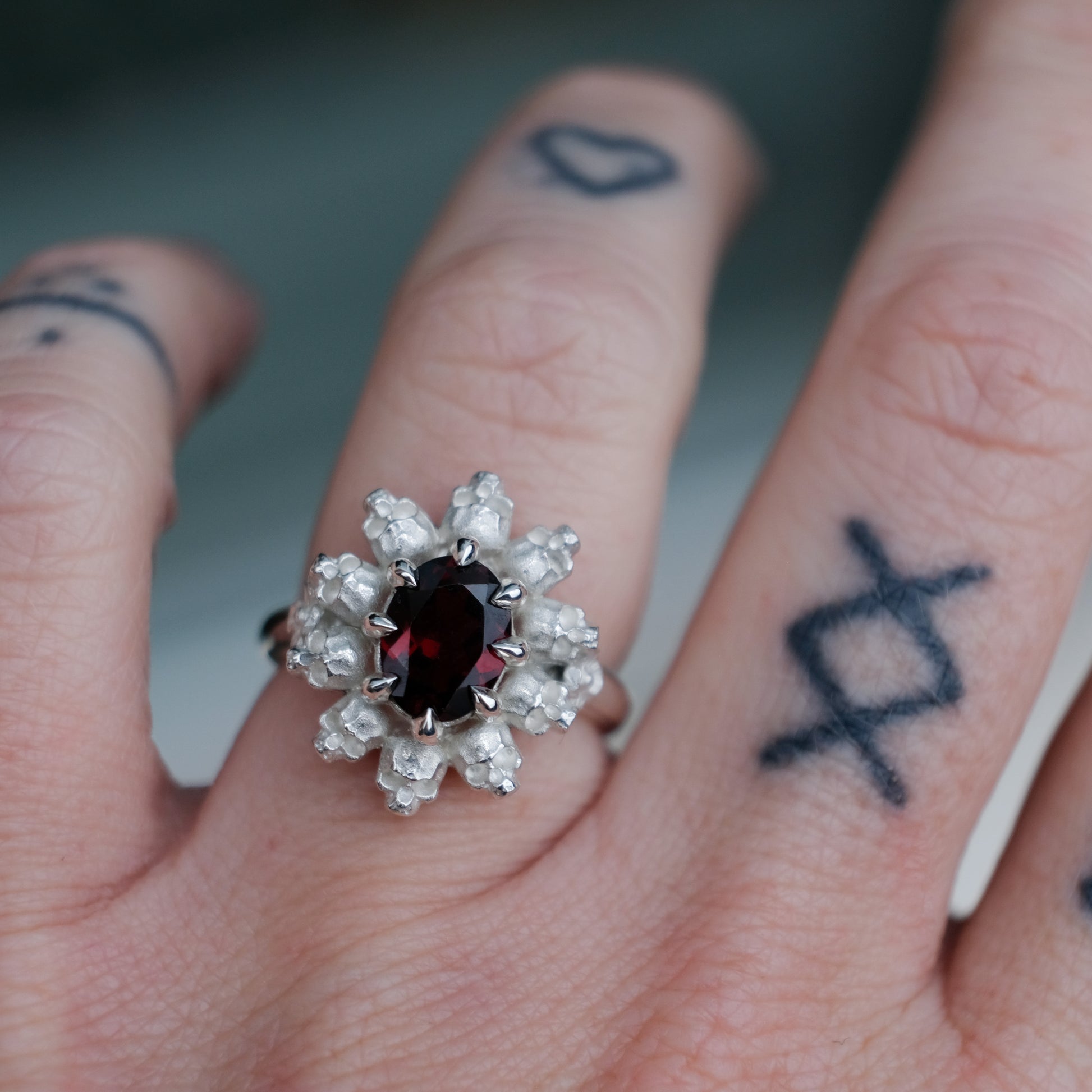 Silver ring with red garnet stone in skull halo design on worn on hand with tattoos, handmade in London, UK