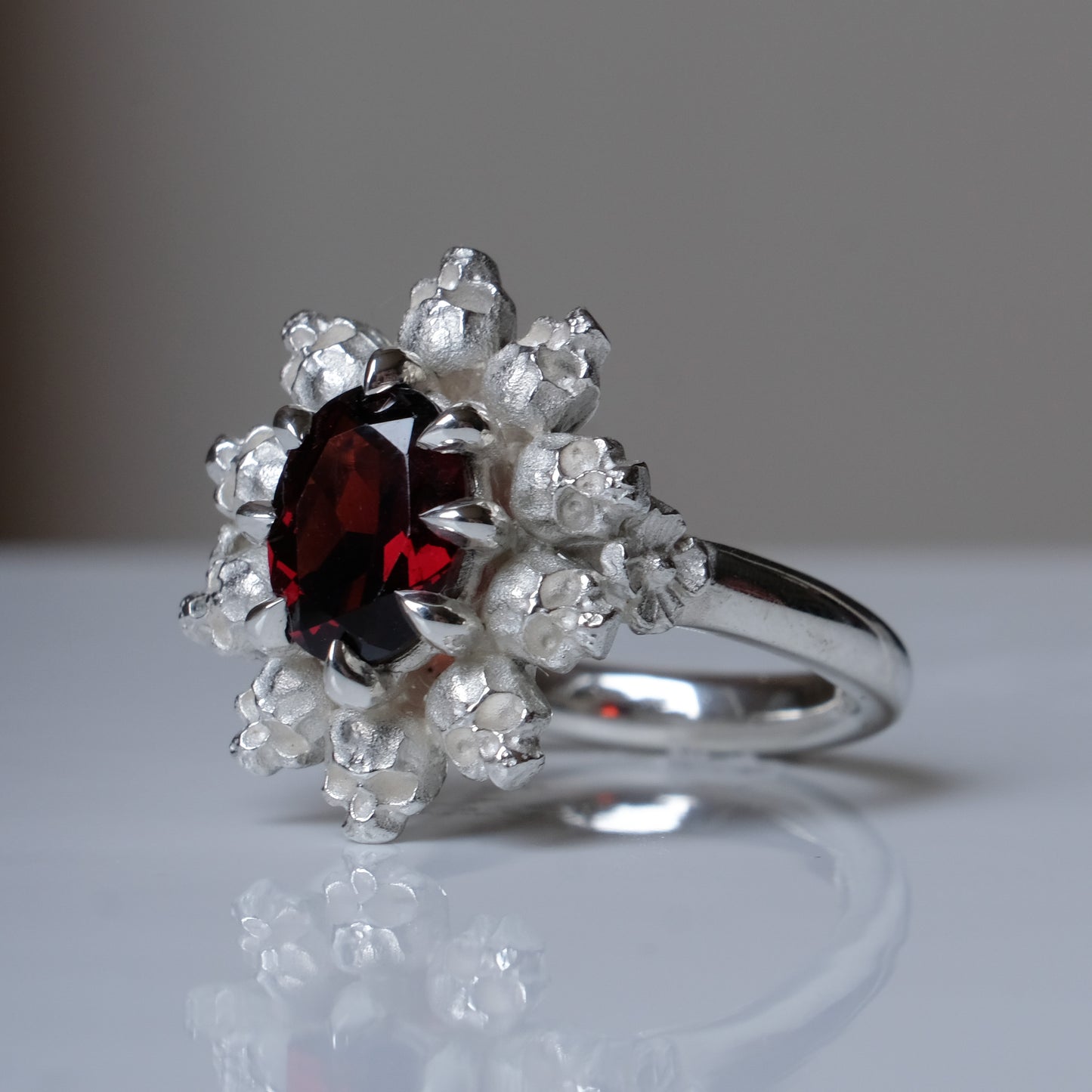 Silver ring with red garnet stone in skull halo design on reflective background handmade in London, UK