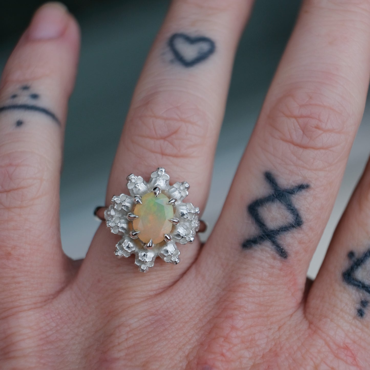 Silver ring with white opal gemstone in skull halo design worn on hand with tattoos handmade in London, UK