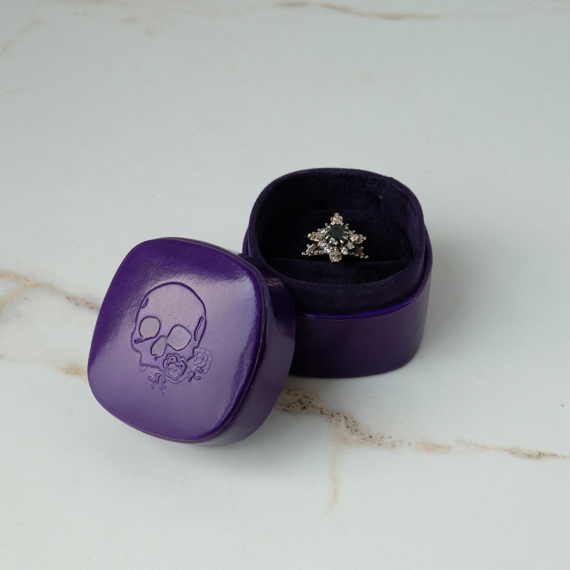 A purple leather engagement ring box with embossed skull logo with skull ring inside by Charlie Shires