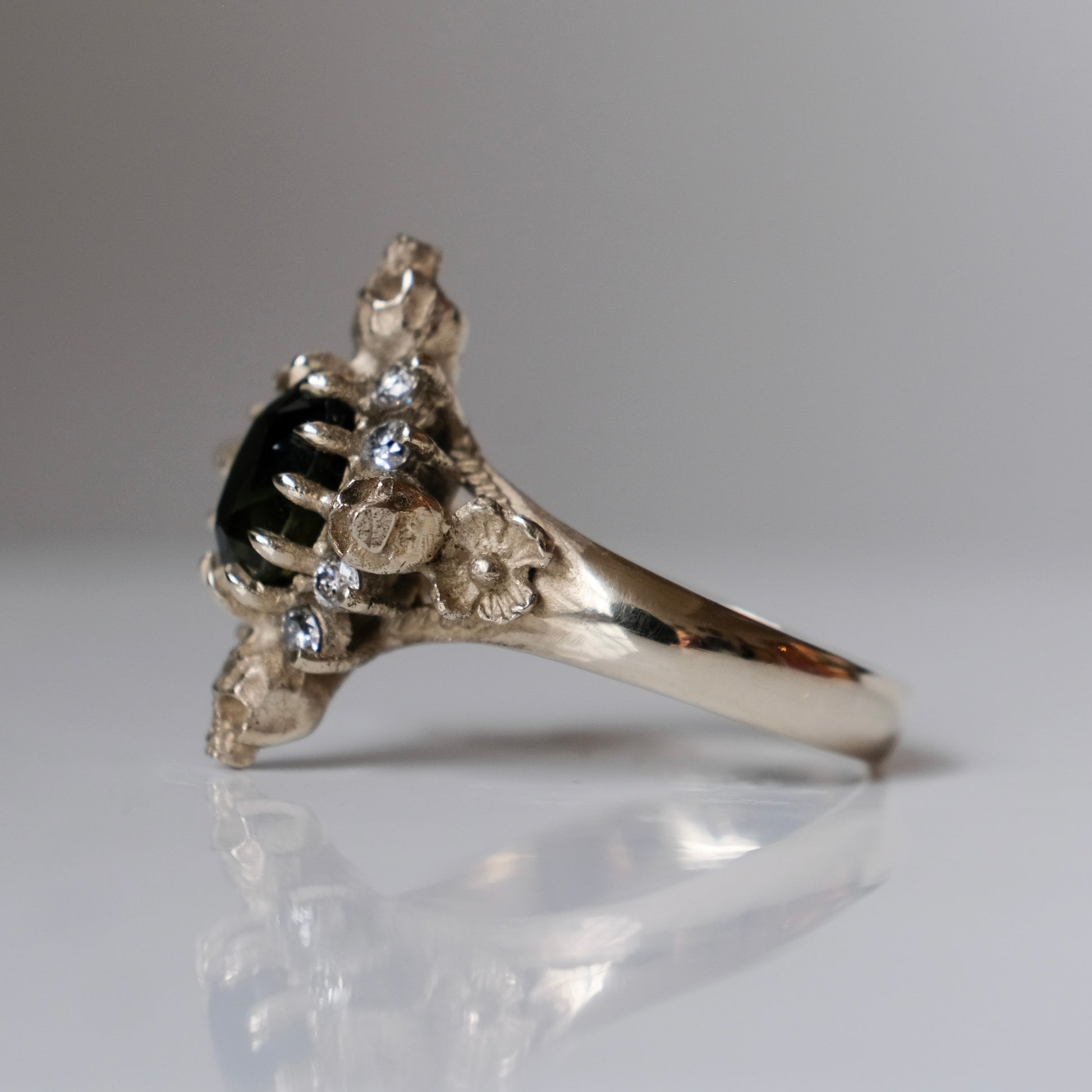 14ct white gold parti sapphire ring with halo of diamonds and skulls. Flowers flank the side of the band. Handmade in London by Charlie Shires