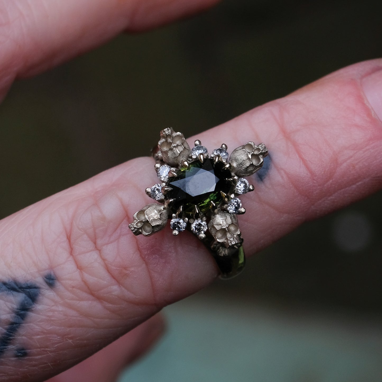 14ct white gold parti sapphire ring with halo of diamonds and skulls. Flowers flank the side of the band. Handmade in London by Charlie Shires