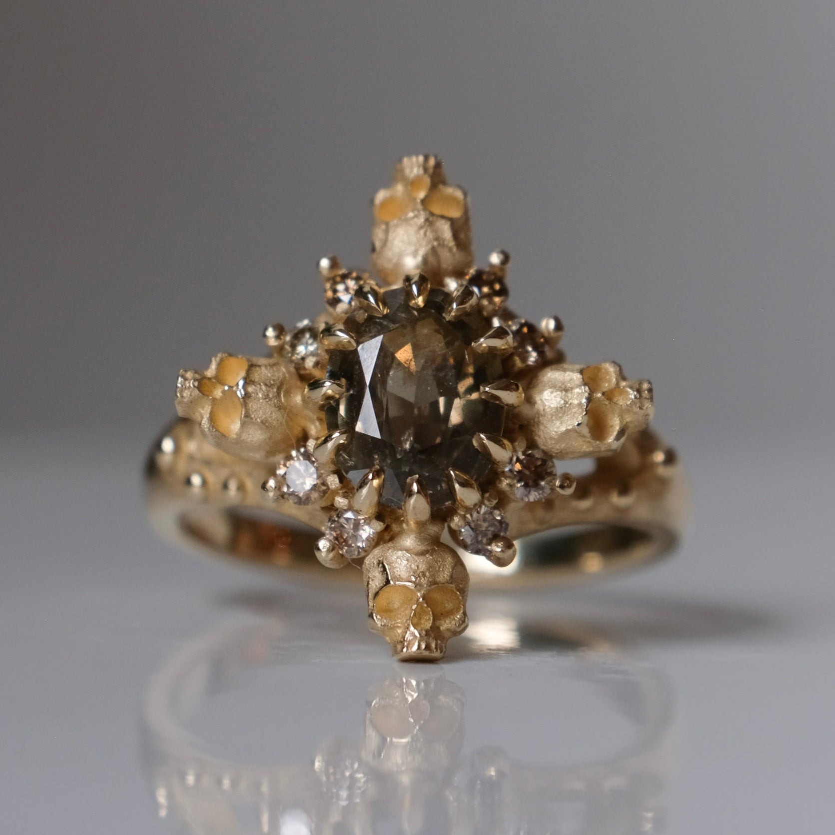 9ct yellow gold engagement ring with unheated sapphire and champagne diamond halo decorated with 4 skulls. 
Handmade in London by Charlie Shires