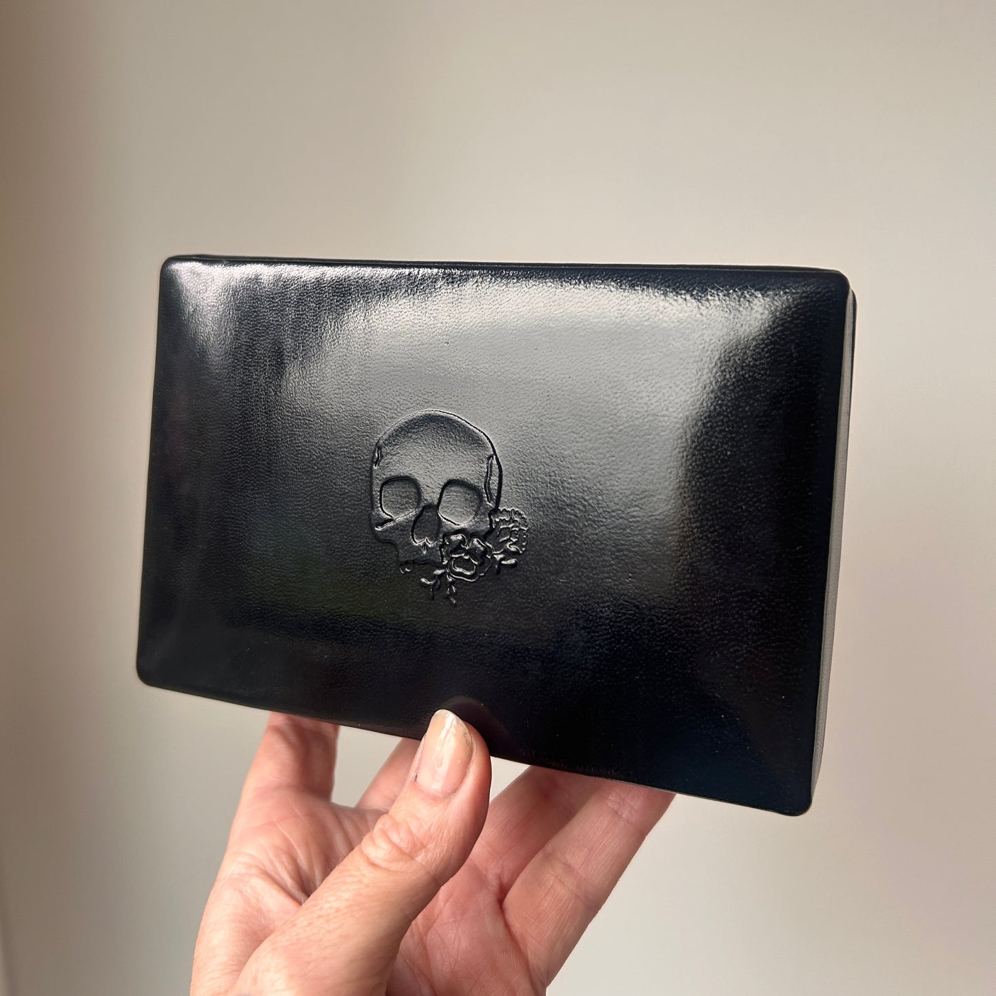 A hand holding a glossy black leather rectangular jewellery box with a raised skull and floral emblem on the front, set against a plain light beige background.