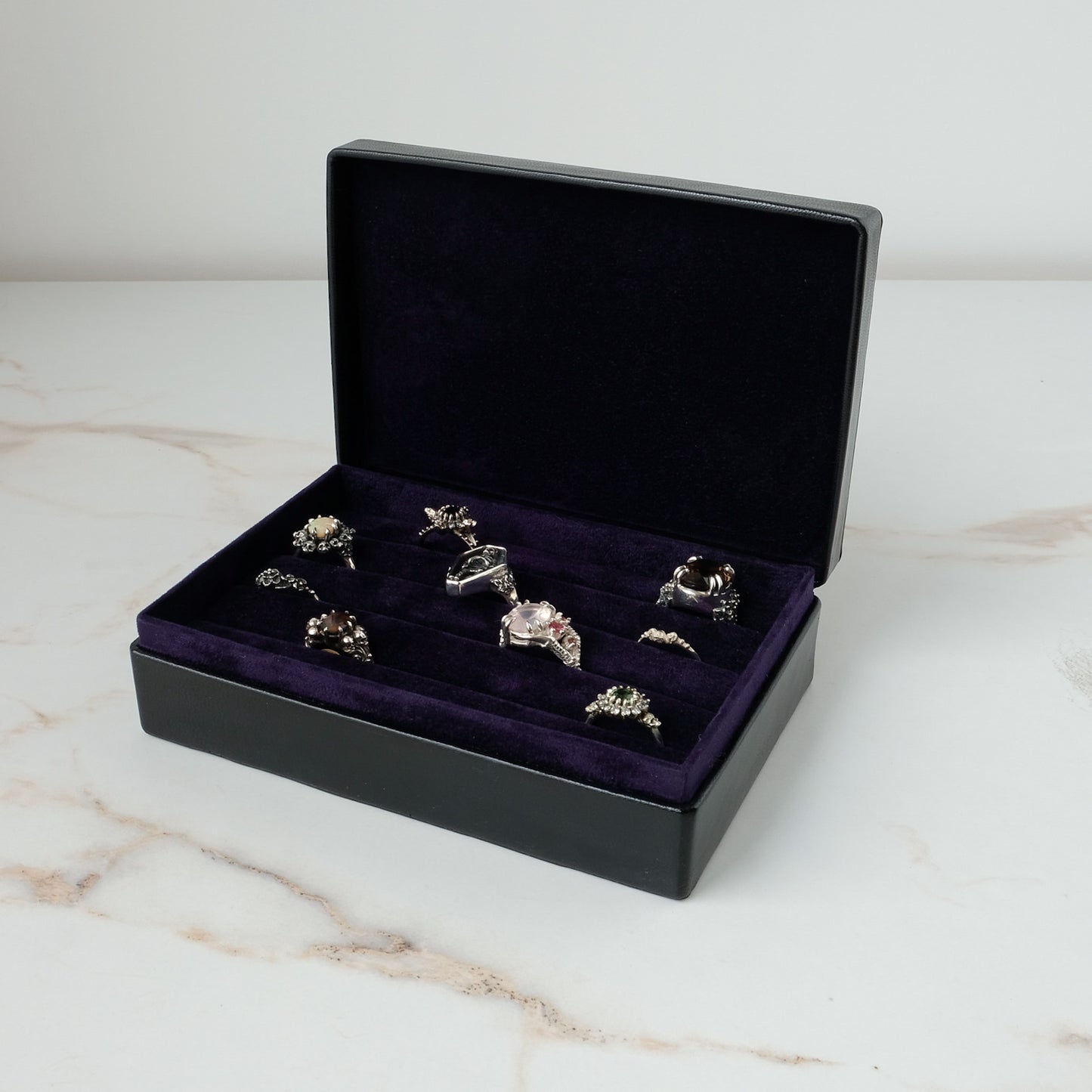 A black leather jewelry ring box with an open lid, lined with deep purple velvet and holding several silver-toned rings, set on a light marble surface against a white background by Charlie Shires