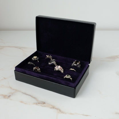 A black leather jewelry ring box with an open lid, lined with deep purple velvet and holding several silver-toned rings, set on a light marble surface against a white background by Charlie Shires