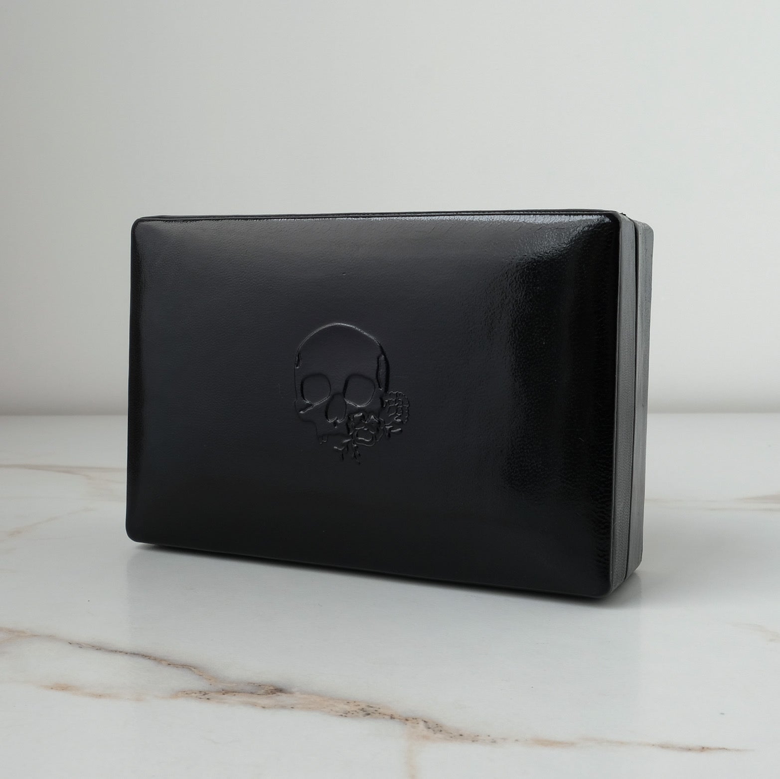 A black leather rectangular jewellery ring box with an embossed skull design on the front, displayed on a white marble surface against a light gray background by Charlie Shires