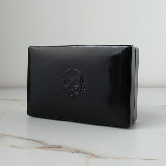 A black leather rectangular jewellery ring box with an embossed skull design on the front, displayed on a white marble surface against a light gray background by Charlie Shires