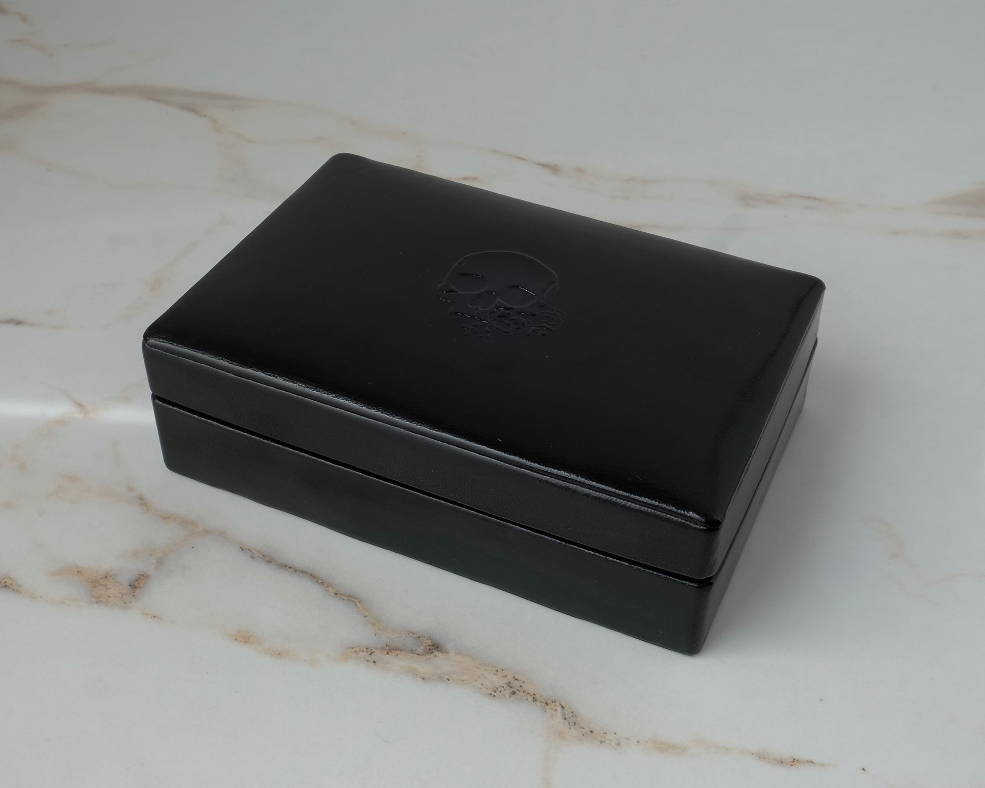 A black rectangular leather jewellery ring box with a debossed skull logo on the lid, resting on a white marble surface by Charlie Shires