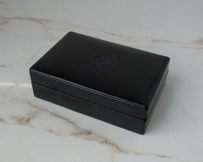 A black rectangular leather jewellery ring box with a debossed skull logo on the lid, resting on a white marble surface by Charlie Shires