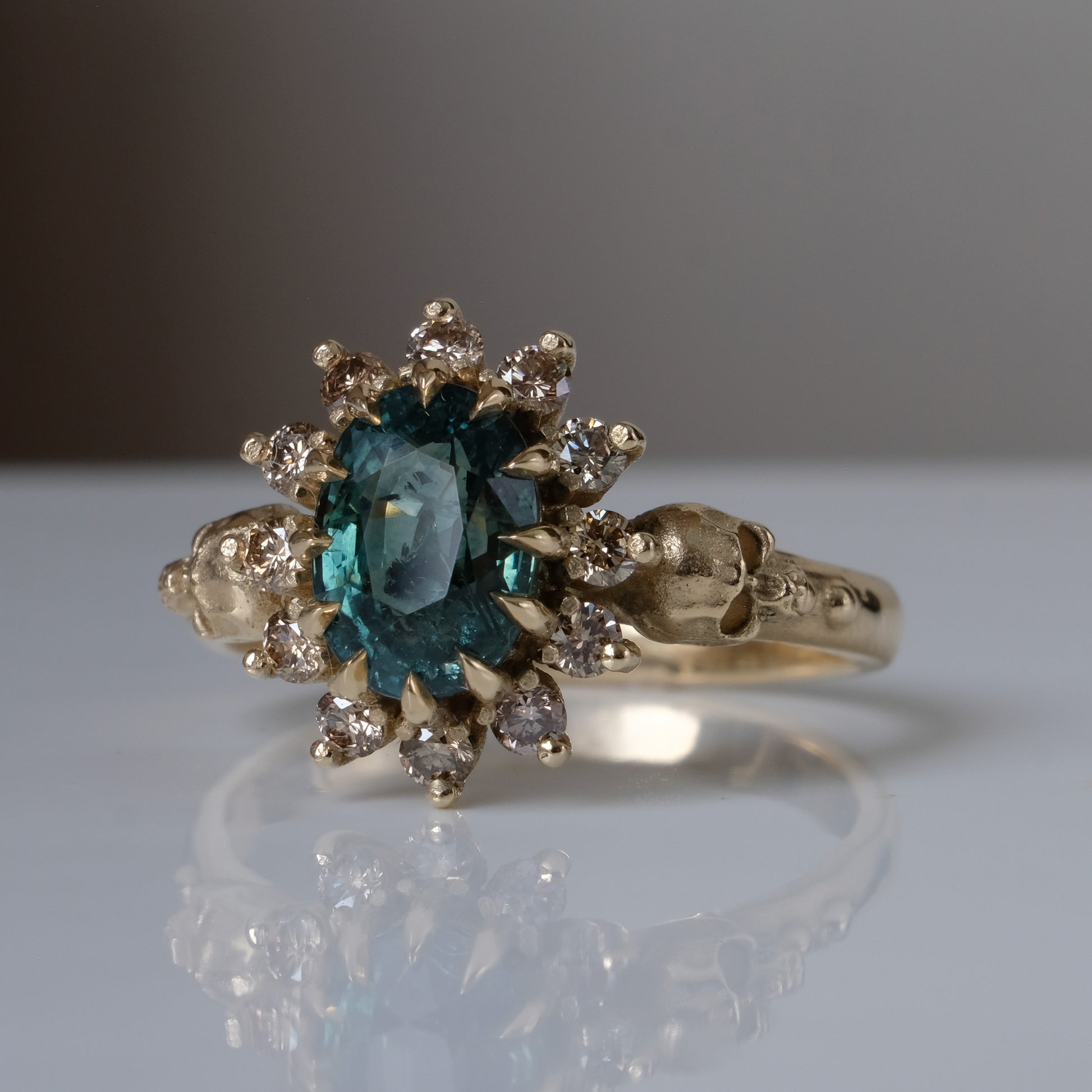 Montana sapphire ring with champagne diamonds in a halo/cluster setting, flanked by two skulls. Handmade in 9ct yellow gold on a white background. Handmade in London from recycled metals.