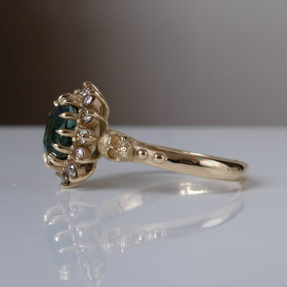 Montana sapphire ring with champagne diamonds in a halo/cluster setting, flanked by two skulls. Handmade in 9ct yellow gold on a white background. Handmade in London by Charlie Shires from recycled metals.