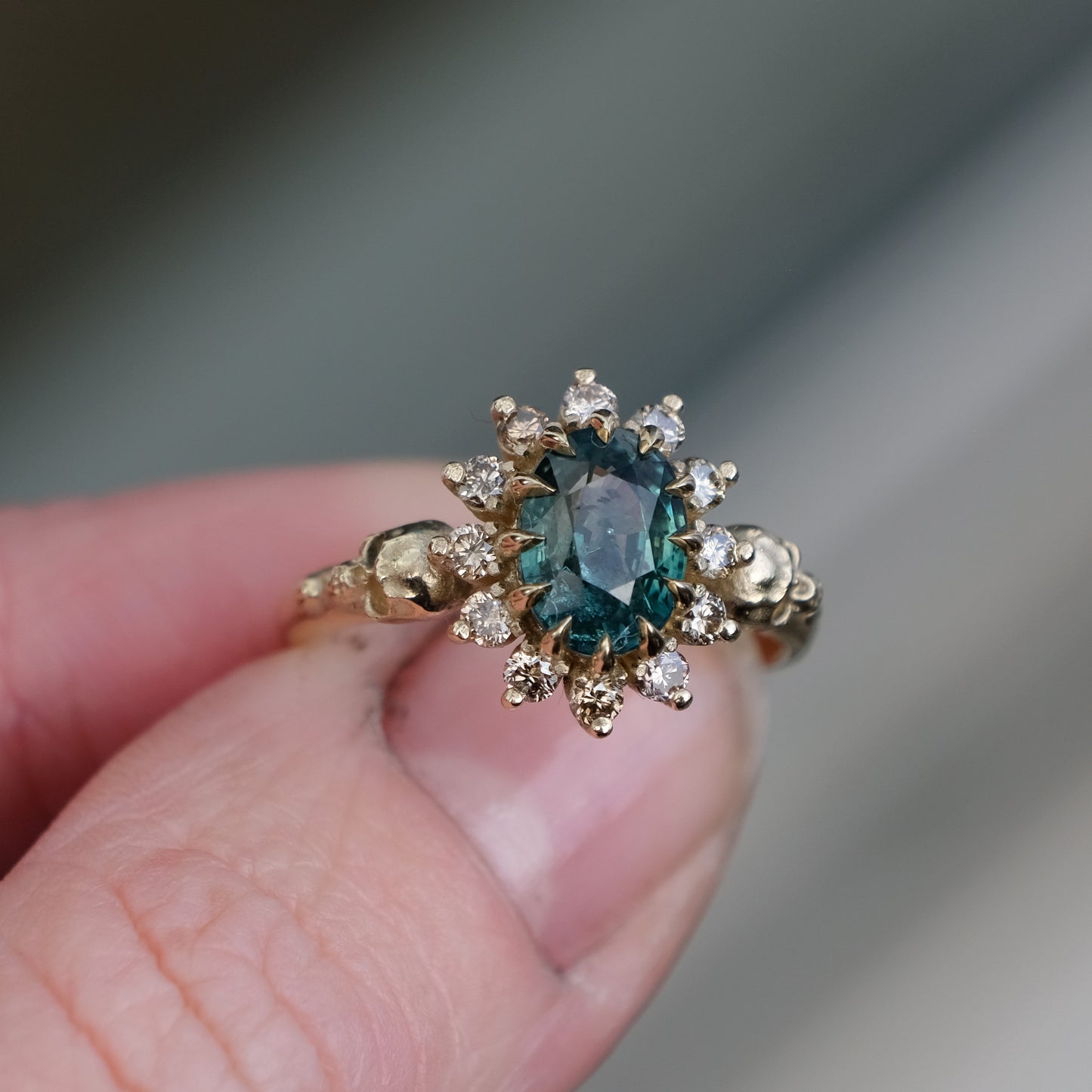 Montana sapphire ring with champagne diamonds in a halo/cluster setting, flanked by two skulls. Handmade in 9ct yellow gold on a white background. Handmade in London by Charlie Shires from recycled metals.