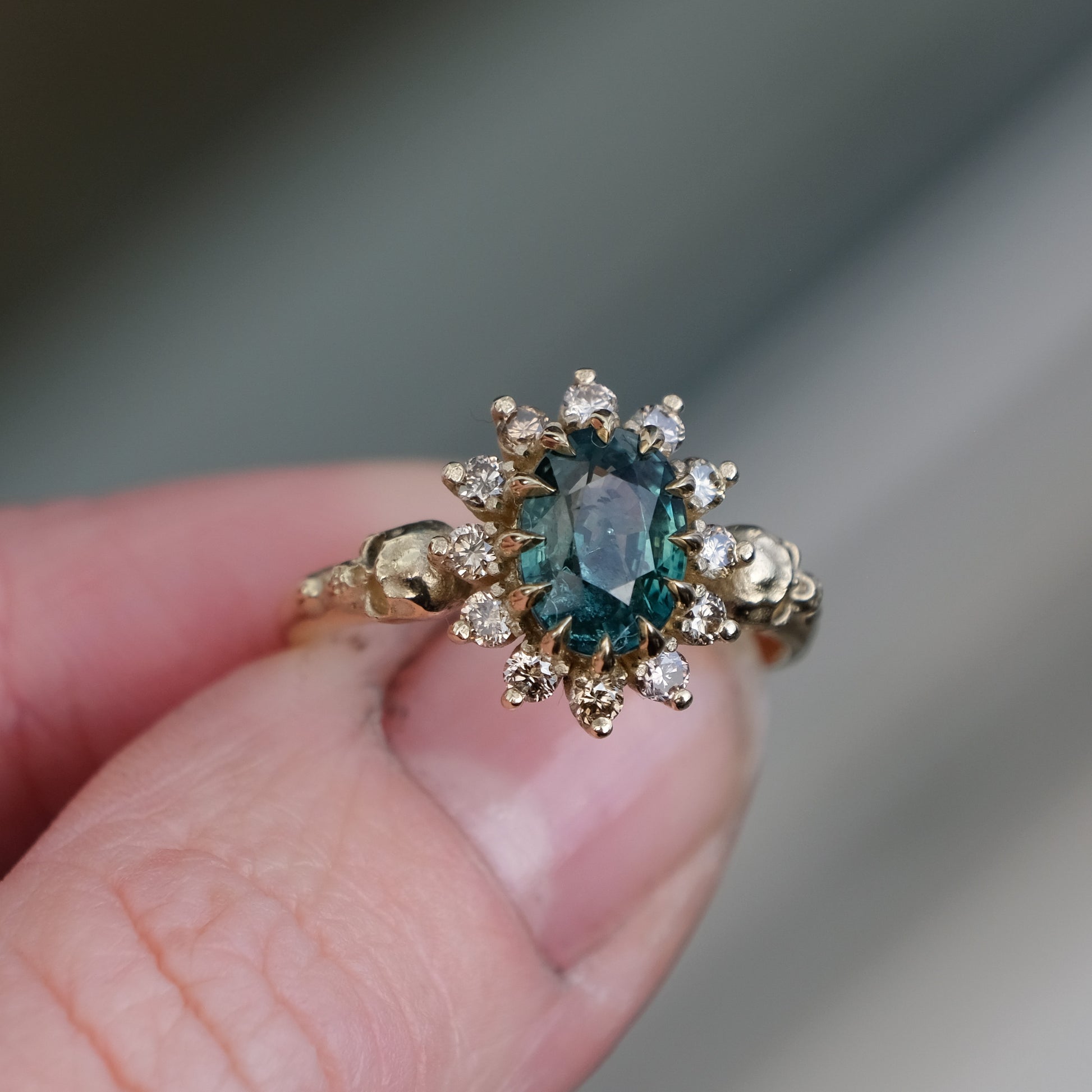 Montana sapphire ring with champagne diamonds in a halo/cluster setting, flanked by two skulls. Handmade in 9ct yellow gold on a white background. Handmade in London by Charlie Shires from recycled metals.