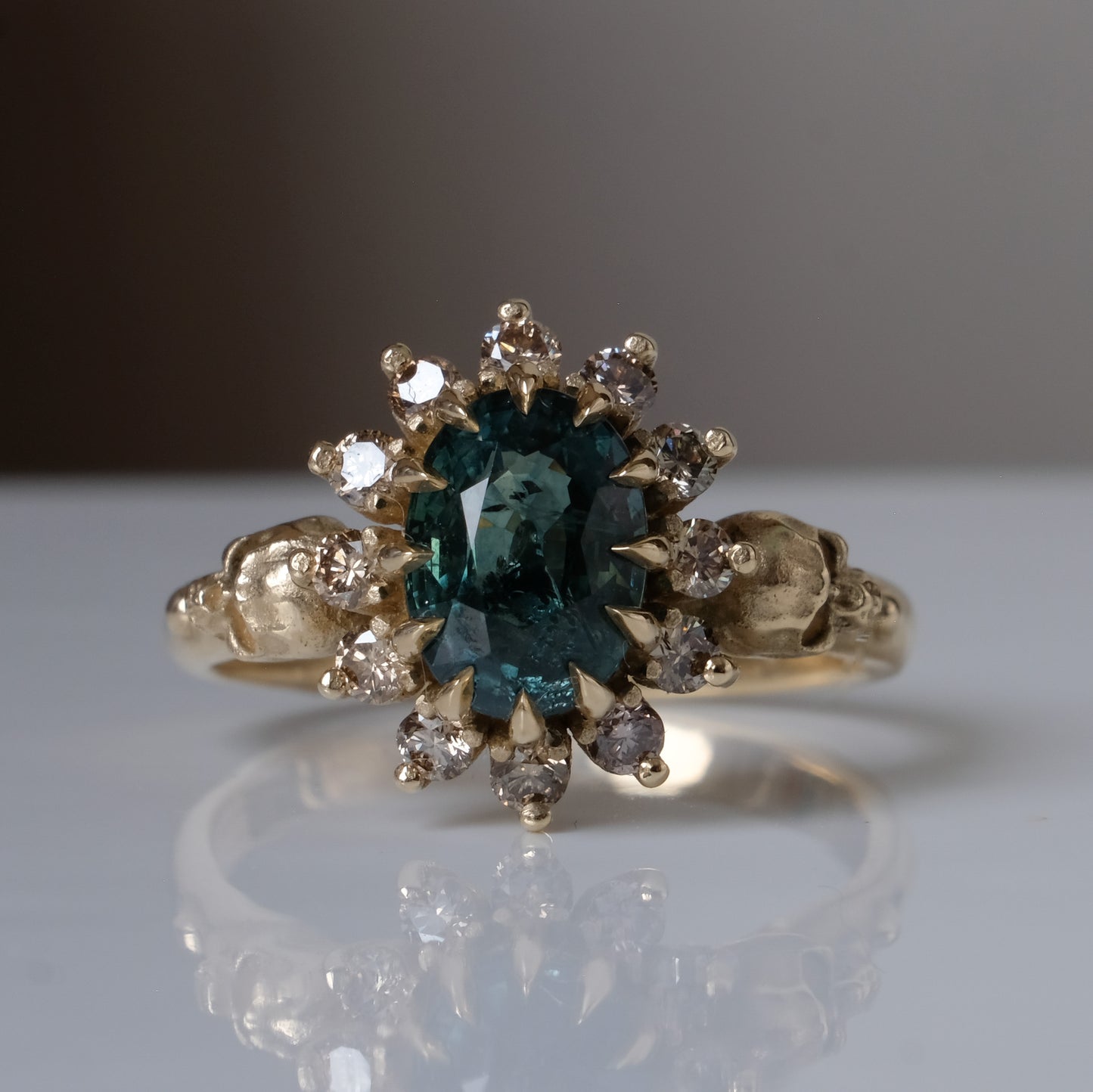 Montana sapphire ring with champagne diamonds in a halo/cluster setting, flanked by two skulls. Handmade in 9ct yellow gold on a white background. Handmade in London from recycled metals.