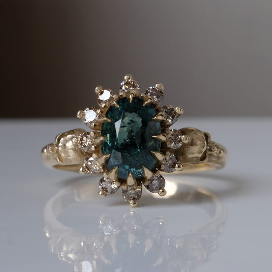 Montana sapphire ring with champagne diamonds in a halo/cluster setting, flanked by two skulls. Handmade in 9ct yellow gold on a white background. Handmade in London from recycled metals.