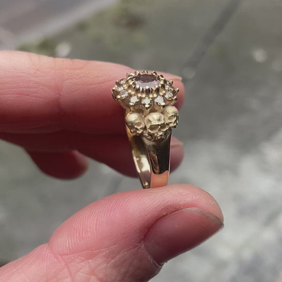 9ct yellow gold engagement ring with central colour change garnet and a champagne diamond halo. 6 skulls flank the side of the setting. 
Handmade in London by Charlie Shires